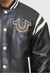 Juodas dirbtinės odos bomberis su baltais dryžiais, mygtukais, ir išsiuvinėtu "True Religion" logotipu auksine ir balta spalva krūtinėje.
