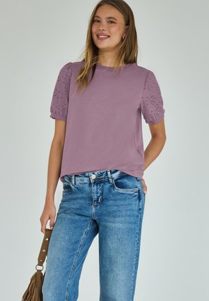 Junge Frau lächelt, trägt ein lavendelfarbenes Hemd mit Lochstick-Puffärmeln und blaue Jeans, hält eine braune Tasseltasche an ihrer Seite.