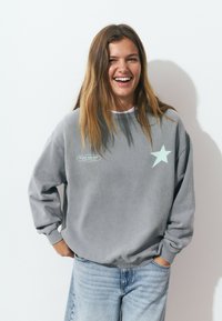 Sweatshirt gris avec un motif d'étoile bleu clair sur le devant, avec des poignets côtelés et un col rond, associé à un jean en denim clair.