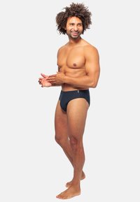 Slip da bagno a righe bianche e nere con un design a vita bassa, realizzati in tessuto morbido, mostrati su un modello maschile in posizione eretta.