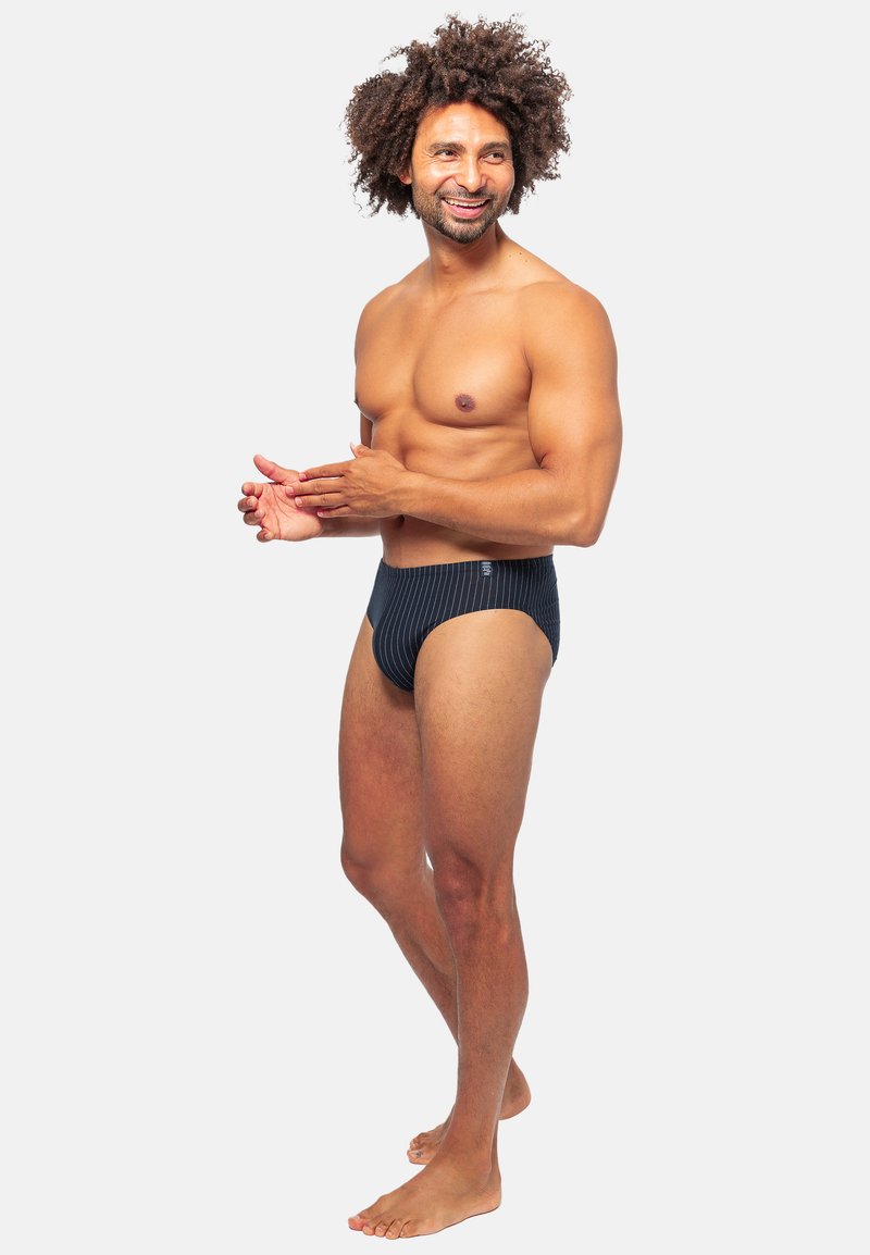 Slip da bagno a righe bianche e nere con un design a vita bassa, realizzati in tessuto morbido, mostrati su un modello maschile in posizione eretta.