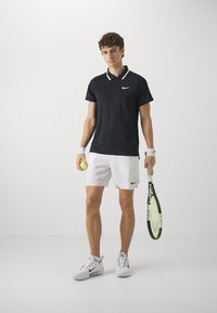Juodas Nike polo marškinėliai su baltais akcentais, balti šortai ir balti sportbačiai. Laikantis geltoną teniso kamuoliuką ir raketę su neono detalėmis.