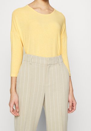 Personne portant un haut jaune ample à manches longues et un pantalon beige taille haute avec des rayures blanches verticales.