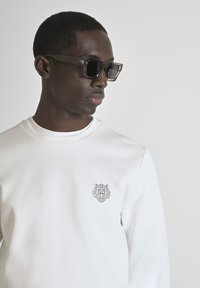 Sudadera blanca con cuello redondo y un emblema de tigre en el pecho; combinada con unas gafas de sol oscuras, que presentan un diseño robusto y geométrico.