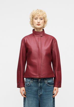 Person mit kurzen lockigen blonden Haaren, die eine rote Lederjacke mit hohem Kragen und locker sitzende blaue Jeans trägt, stehend vor einem schlichten Hintergrund.