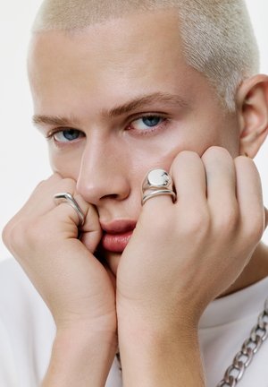 Des bijoux en argent sur des mains, vue de près. La personne a la peau claire, des cheveux courts décolorés, des yeux bleus et des traits doux. Fond blanc uni.