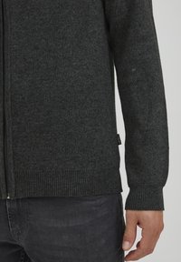 Un sweat à fermeture éclair gris foncé en tissu tricoté doux. Il présente des poignets et un ourlet côtelés, avec une petite étiquette de marque sur le côté.