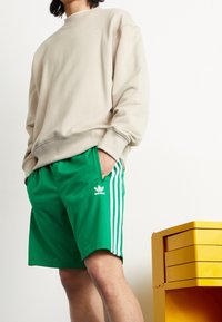 Beige sweatshirt med rund hals, tillsammans med gröna träningsshorts med vita sidostycken och en adidas-logotyp. Gul förvaringslåda i närheten.