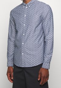 Košile na knoflíky v námořnické modři s bílým geometrickým vzorem, dlouhými rukávy a rozkládacím límcem. Má zakřivený lem a bílé knoflíky.