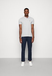 Polo shirt grigio chiaro con colletto e maniche corte, abbinato a pantaloni blu navy e sneakers bianche. Presenta un piccolo logo verde sul petto.