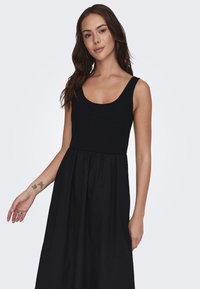 Mustapuku, jossa on ribatusta, vartaloa myötäilevä yläosa ja soljuva hame. Pehmeä kangas, leveät hihnat ja vaatimaton pyöreä pääntie.