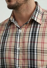 Chemise à carreaux beige clair, avec des rayures rouges, noires et grises. Col boutonné, boutons blancs, en coton, texture visible.