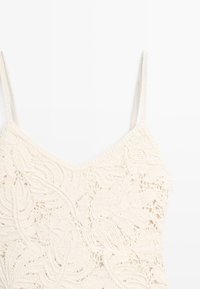 Cremafarbene gehäkelte Camisole mit dünnen Trägern und floralem Spitzenmuster auf weißem Hintergrund.