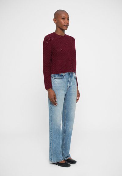Mulher em pé, vestindo um cardigan de malha bordeaux com botões, jeans claros de cintura alta e sapatilhas pretas, em fundo branco liso.