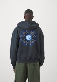 Sweatshirt preto com design gráfico azul nas costas, apresentando um emblema circular rodeado por padrões intricados. Tecido espesso e capuz com cordão.