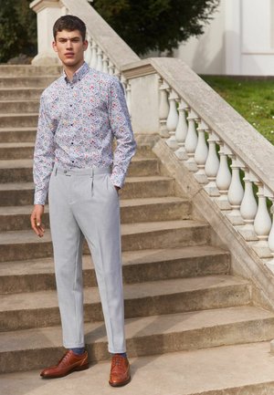 Biała koszula w wielokolorowy kwiatowy wzór, szare dopasowane spodnie i brązowe skórzane buty formalne. Stoi na kamiennych schodach.