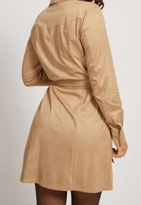 Brązowa zamszowa sukienka koszulowa z długimi rękawami, zapięciem na guziki przy mankietach, pasek w talii oraz rozkloszowanym dołem. Posiada szew z tyłu oraz wykładany kołnierz.