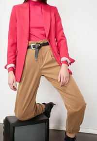 Fuchsiový blazer, růžový rolák s dlouhým rukávem, béžové široké kalhoty, černý opasek se stříbrnou sponou a černé kotníkové boty. Model pózuje na televizoru.