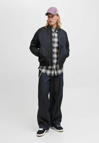 Una chaqueta bomber negra sobre una camisa de cuadros, combinada con jeans de pierna ancha y zapatillas negras. La gorra es de un morado claro con parte trasera de malla.