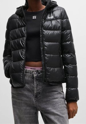 Femme portant une doudoune noire, un haut court noir, un jean gris et une ceinture rose avec la marque "HUGO" visible.