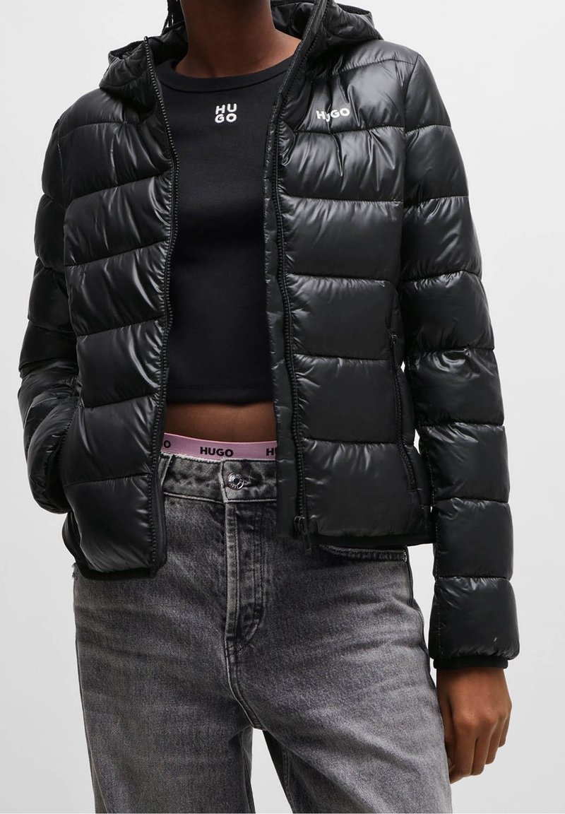 Femme portant une doudoune noire, un haut court noir, un jean gris et une ceinture rose avec la marque "HUGO" visible.