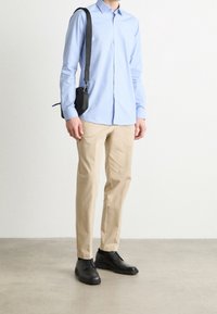 Camicia azzurra chiara a maniche lunghe con bottoni, chinos beige, scarpe stringate nere e una piccola borsa a tracolla nera con cinturino.