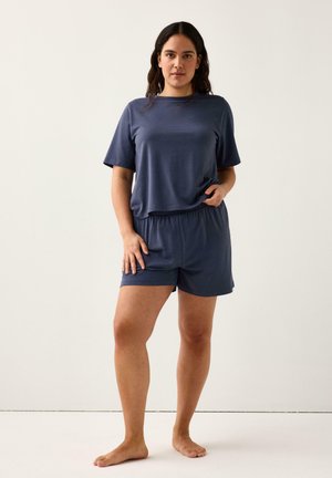 Kvinde stående barfodet iført løs mørkeblå kortærmet skjorte og matchende elastiske taljeshorts mod ensartet lyst baggrund.