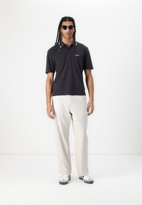 Tricou polo bleumarin cu mâneci scurte, guler contrastant, pantaloni bej deschis și adidași gri. Ochelarii de soare negri adaugă o notă modernă.