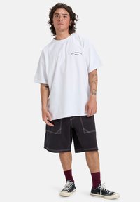 Hvit oversized bomulls t-skjorte med svart logo og korte ermer, kombinert med mørke shorts med kontrastfargede hvite sømmer og lommer.