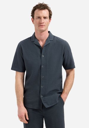 Mann mit lockigem Haar, der ein dunkles, strukturiertes kurzärmliges Hemd mit Knöpfen und passende Hose trägt, mit einer Hand in der Tasche vor weißem Hintergrund.