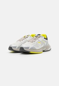 Sneakers bianchi con sovrapposizioni in rete e sintetico, dettagli grigi e una linguetta in rete nera. Presentano un tacco giallo brillante e suola in gomma.
