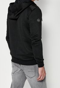 Felpa nera con cappuccio e trama a coste, caratterizzata da eleganti dettagli sulle spalle e una toppa con logo sulla manica. Abbinata a jeans grigi.