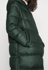 Mørkegrøn dynejakke med hætte, der har kviltede sektioner, sidelommer og en glat, skinnende tekstur. Lynlåsdetalje på siden.