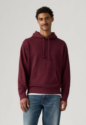 Man met kort krullend haar en snor, die een bordeauxrode hoodie met voorzak en blauwe jeans draagt, staand voor een effen achtergrond.
