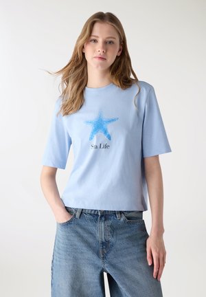 Giovane donna con una maglietta azzurra con una stella marina blu e la scritta "Sea Life", abbinata a jeans larghi blu, in piedi con una mano in tasca.