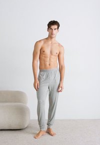 Pantaloni de pijama gri, cu croială lejeră, talie elastică, manșete tapered și o textură moale. Purtați fără cămașă, stând pe un covor deschis.