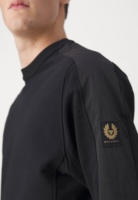 Svart tröja med texturerade ärmar och en logotyp på vänster axel; tillverkad av ett mjukt, elastiskt material för komfort.