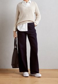 Beige gebreide trui, wit overhemd met kraag eronder, donkere wijde broek, witte sneakers, een donkerkleurige totebag vasthoudend, houten vloer.