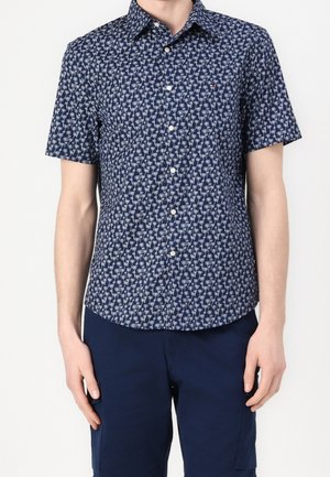 Mann trägt ein marineblaues Kurzarmhemd mit weißen Palmenmuster und marineblaue Hose, steht vor einfachem Hintergrund.