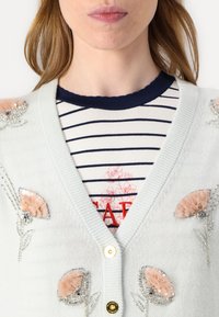Cárdigan azul claro con bordado floral en rosa y plata. Presenta un escote en V, botones y una camiseta a rayas en capas debajo.