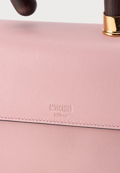 Gros plan d’un sac à main en cuir rose avec le logo Moschino Milano embossé sur le rabat et deux poignées arrondies marron foncé avec des accents dorés.