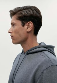 Sivá mikina s kapucňou a sťahovacou šnúrkou, s jemnou textúrou a diskrétnymi prešitými akcentmi na pleciach. Bočný profil modela.