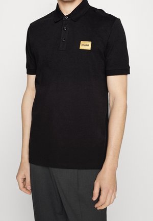 Polo negro hecho de tela lisa, con cuello acanalado, tapeta de tres botones y un parche rectangular con logo dorado en el pecho.