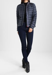 Donna che indossa una giacca trapuntata blu navy sopra una maglietta nera, jeans skinny scuri e stivaletti neri, in piedi su uno sfondo chiaro e uniforme.