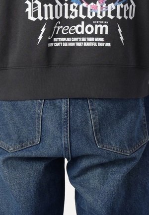 Czarna bluza z białym napisem "Undiscovered freedom" oraz cytatem z motylem, nad niebieskimi dżinsami z tylnymi kieszeniami.