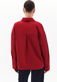 Camicia rossa a maniche lunghe con colletto, caratterizzata da una piega centrale sul retro e polsini con bottoni. Tessuto liscio e vestibilità leggermente over.
