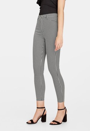 Femme portant un pantalon ajusté à motif pied-de-poule noir et blanc et des sandales noires à talons ouvertes, debout devant un fond blanc.