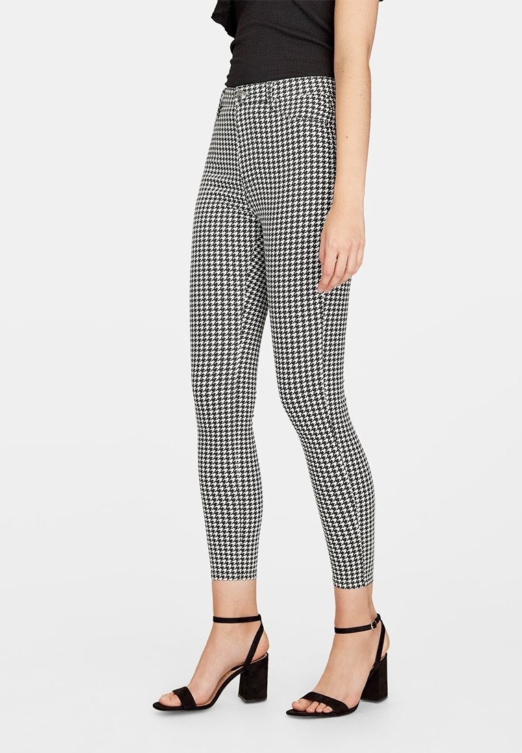 Femme portant un pantalon ajusté à motif pied-de-poule noir et blanc et des sandales noires à talons ouvertes, debout devant un fond blanc.