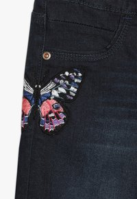 Tmavé džíny z denimu s velkou, vícebarevně vyšitou motýlicí na levém stehně. Motýl obsahuje růžové, modré a bílé detaily.