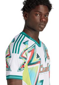 Junger Mann mit kurzen, lockigen Haaren, der ein buntes Sporttrikot mit geometrischen Mustern und grünen Schulterstreifen trägt und in die Kamera schaut.
